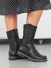 Black leather boots with inner zippers on feet, standing on gray floor, paired with a black and white polka dot skirt. The boots have sturdy soles and a sleek design