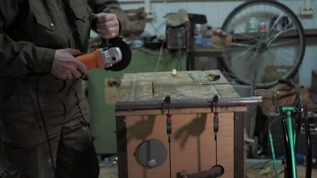 Workshop. A worker uses a angle grinder to cut a steel pipe that is fixed to a workbench with clamps along a drawn line in front of a bicycle.