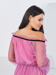 Brunette woman in a vibrant off-shoulder pink dress with sheer sleeves, facing away, smiling...