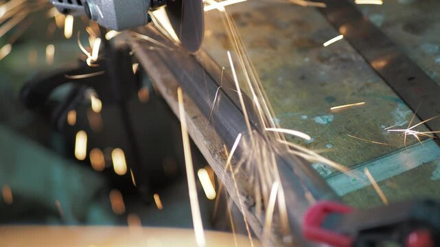 Workshop. A worker uses a angle grinder to cut a steel pipe that is fixed to a workbench with a clamp along a marked line. Close-up.