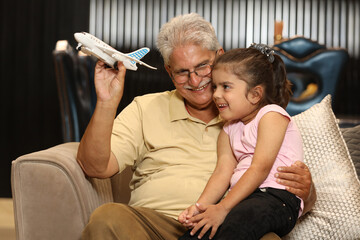 Indian happy grand father and grand daughter playing airplane while sitting on sofa.