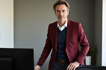 Confident stylish middle-aged businessman or creative director in a maroon blazer standing at his desk in a modern office, looking at camera with a powerful, serious gaze