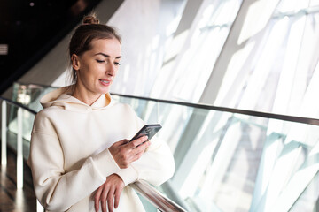 Smiling young woman looking at phone while standing near panoramic window