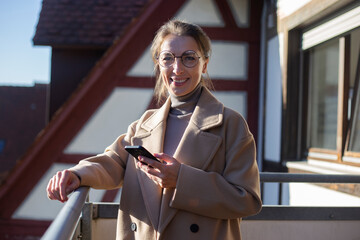 Smiling young woman in coat with phone standing on terrace