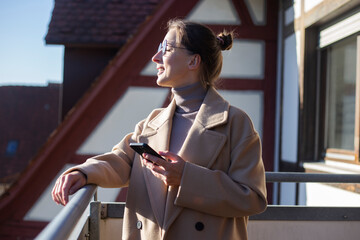 Young woman in a coat with a phone standing on the terrace