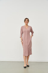 Smiling woman stands wearing a knee-length mauve dress with three-quarter sleeves. She holds her...