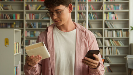 Male student man guy teenager in library choosing between book and smartphone dropping copybook...