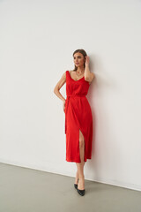 Red sleeveless dress with a belt accentuates the figure of a woman leaning against a white wall....