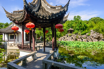 A traditional Chinese garden with winding bridge leading to a pavilion, adorned with red lanterns...