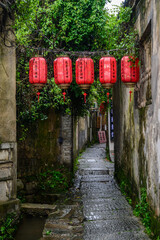 Lush greenery drapes ancient stone walls framing a narrow alley, adorned with red lanterns bearing...