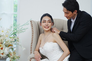 Asian groom in black suit with diamond necklace for beautiful Asian bride in white dress sitting on...