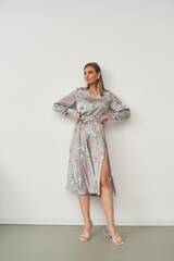 Woman in floral-patterned gray dress standing against a plain wall with hands on hips, looking to...
