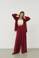 Young woman in a burgundy pantsuit with wide-leg trousers, wearing a white crop top underneath. She...