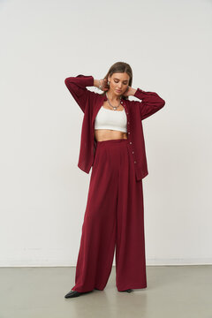 Young woman in a burgundy pantsuit with wide-leg trousers, wearing a white crop top underneath. She adjusts her hair with both hands, standing confidently on a beige floor