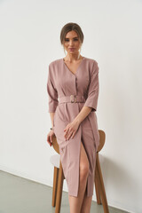 Elegant woman in mauve knee-length dress with a belt stands leaning on a wooden chair, hands...