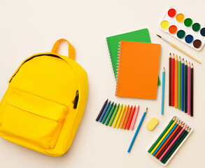 Flat lay of colorful school and art supplies on a beige background. A bright yellow backpack is surrounded by notebooks, colored pencils, crayons, watercolor paint set, paintbrush, and an eraser. Crea