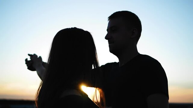 Two lovers, a guy and a girl, dancing at sunset on a high roof. Their dancing silhouettes are visible
