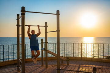 A senior man does pull-ups on a bar by the sea at sunrise. His silhouette highlights calmness, vitality, and mindful aging. Golden backlight enhances the peaceful morning atmosphere.