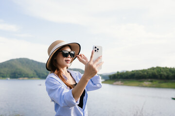 A young Asian woman leans on a white car, gazing thoughtfully at a serene mountain lake, embracing...