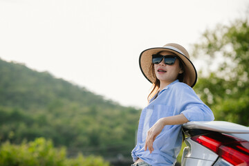 A young Asian woman leans on a white car, gazing thoughtfully at a serene mountain lake, embracing...