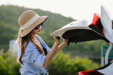 A young Asian woman leans on a white car, gazing thoughtfully at a serene mountain lake, embracing...