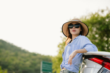 A young Asian woman leans on a white car, gazing thoughtfully at a serene mountain lake, embracing...