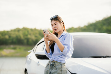A young Asian woman leans on a white car, gazing thoughtfully at a serene mountain lake, embracing...