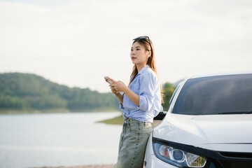 A young Asian woman leans on a white car, gazing thoughtfully at a serene mountain lake, embracing...