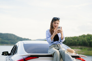 A young Asian woman leans on a white car, gazing thoughtfully at a serene mountain lake, embracing...