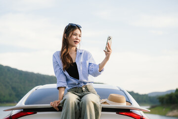 A young Asian woman leans on a white car, gazing thoughtfully at a serene mountain lake, embracing...