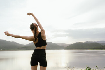 Obraz premium A young Asian woman practices stretching or yoga outdoors by a serene lake with mountain views, embracing fitness, calmness, and a healthy lifestyle in nature’s tranquility.