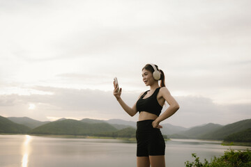 A young Asian woman practices stretching or yoga outdoors by a serene lake with mountain views, embracing fitness, calmness, and a healthy lifestyle in nature&rsquo;s tranquility.