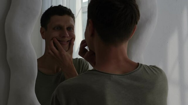 Young man forcing a smile in the mirror. Concept of depression, mental health struggles, and emotional pain. Minimalist interior with soft light.