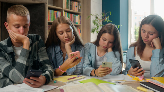 Four Caucasian pupils classmates friends teenagers one boy guy with three girls using mobile phones gadgets addict internet addiction smartphones overuse dependency in library study problem school