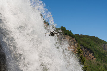 A modern quadcopter with a camera flies in front of a powerful waterfall, capturing a scenic...
