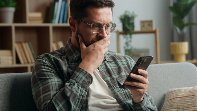 Caucasian adult man male guy at home living room nervous looking at mobile phone bad news amazed shocked close mouth with hand message from bank stressed smartphone notification online error scam - Powered by Adobe