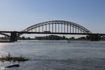 The iconic Waalbrug, a historic arch bridge, spans the Waal River in Nijmegen under a bright sky, symbolizing strength and connection in the Dutch landscape.