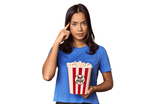 Young woman with popcorn bucket in studio pointing temple with finger, thinking, focused on a task.