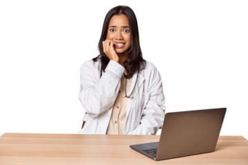 Young Filipino doctor with laptop in studio biting fingernails, nervous and very anxious.