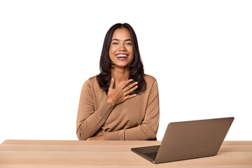 Filipino woman at desk with laptop laughs out loudly keeping hand on chest.