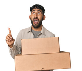 Hispanic male holding cardboard boxes in studio pointing upside with opened mouth.