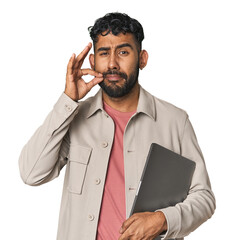 Hispanic male holding laptop in studio with fingers on lips keeping a secret.