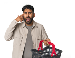 Hispanic male holding shopping basket in studio showing a disappointment gesture with forefinger.