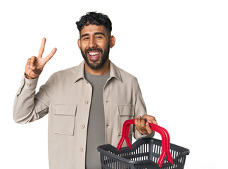Hispanic male holding shopping basket in studio showing number two with fingers.