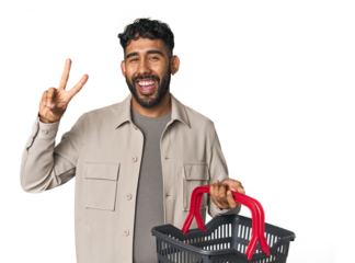 Hispanic male holding shopping basket in studio showing number two with fingers.