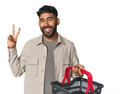 Hispanic male holding shopping basket in studio joyful and carefree showing a peace symbol with fingers.