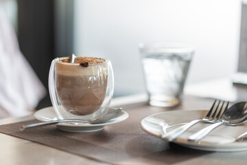 transparent glass mug of hot chocolate on top with foam and dark chocolate flakes,being served dining table,refreshment drink during the day
