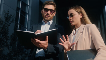 Caucasian two coworkers colleagues businessman business male guy man with businesswoman girl female woman in sunglasses style talking discuss corporate papers documents project holding folders in city