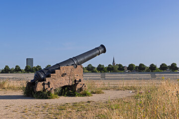 An historic black cannon, a remnant of past military might, stands in a dry, grassy field under a clear blue sky, with a distant church spire and modern buildings.