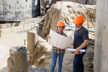 Two construction engineers in hard hats review building plans at an active construction site. An architect and a worker discuss project details amidst a concrete construction background.